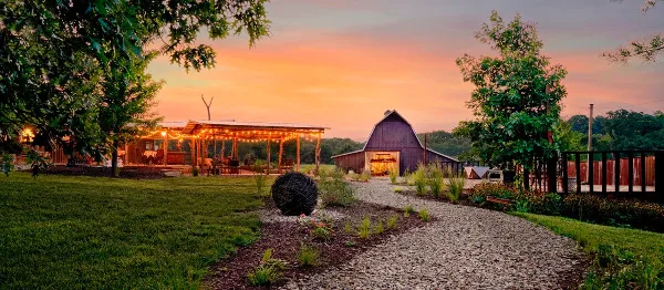 A wide shot of a country wedding venue at sunset featuring a gravel path, a covered pavilion with string lights, and a large barn.