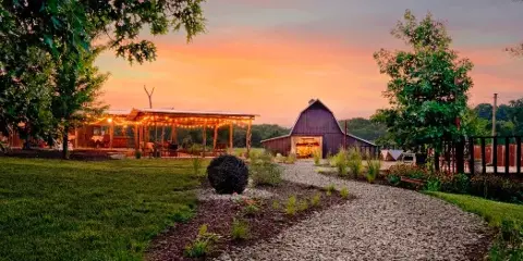 A wide shot of a country wedding venue at sunset featuring a gravel path, a covered pavilion with string lights, and a large barn.