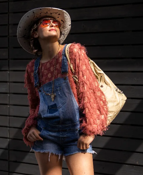 A fashion portrait of a woman wearing a wide-brimmed hat, sunglasses, and denim overalls, standing against a dark wooden slat wall.