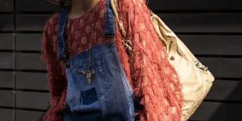 A fashion portrait of a woman wearing a wide-brimmed hat, sunglasses, and denim overalls, standing against a dark wooden slat wall.