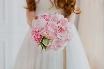 A bride in a white wedding dress holding a round bouquet of pink peonies.