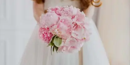 A bride in a white wedding dress holding a round bouquet of pink peonies.