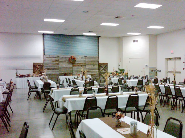 Indoor event hall with long tables covered in white linens, chairs, and a rustic wooden backdrop.