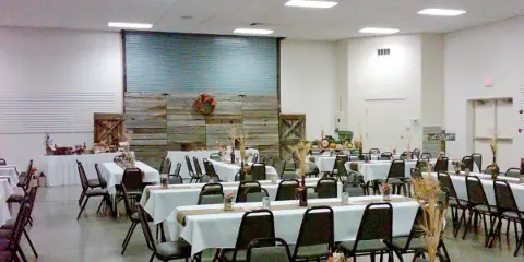 Indoor event hall with long tables covered in white linens, chairs, and a rustic wooden backdrop.