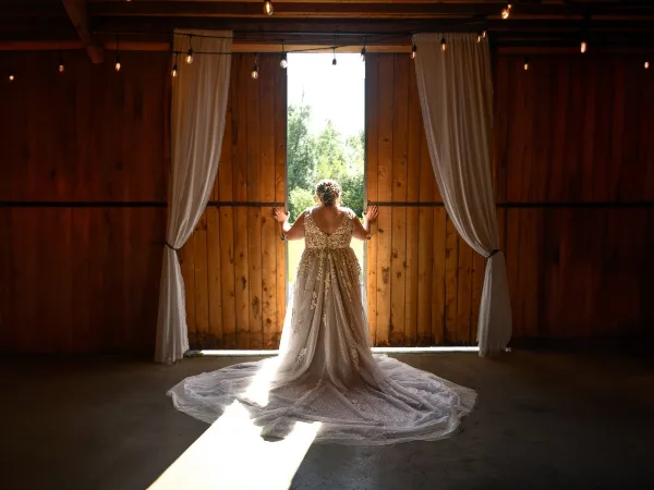 Bride in an embroidered gown standing with back to camera at the open doorway of a wooden barn with draped curtains and string lights.
