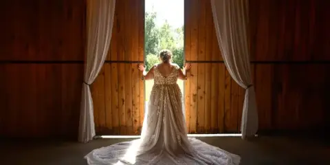 Bride in an embroidered gown standing with back to camera at the open doorway of a wooden barn with draped curtains and string lights.