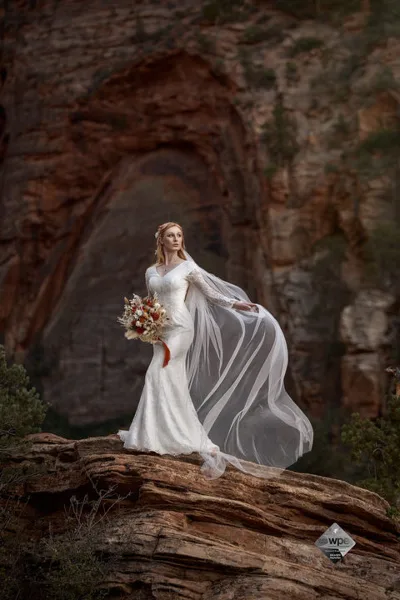 A bride in a white wedding dress and veil stands on a rocky outcrop in a red rock canyon, holding a bouquet.
