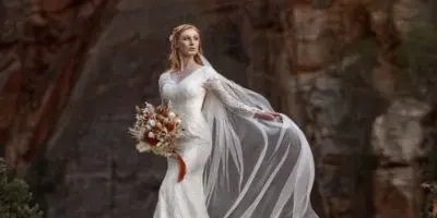 A bride in a white wedding dress and veil stands on a rocky outcrop in a red rock canyon, holding a bouquet.