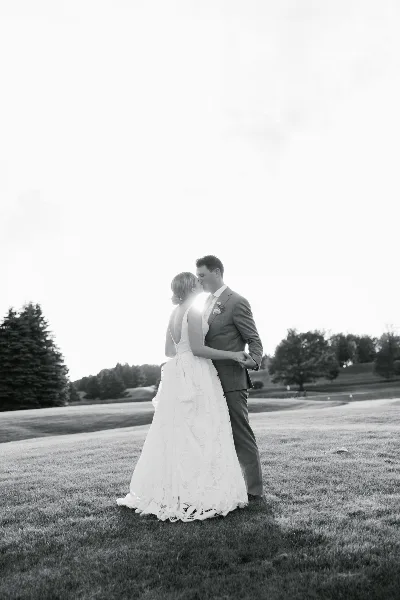 A black and white photograph of a bride and groom kissing on a grassy lawn with trees in the background.