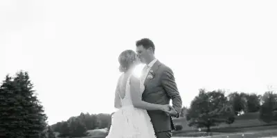 A black and white photograph of a bride and groom kissing on a grassy lawn with trees in the background.
