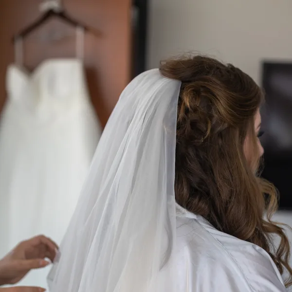A bride with a styled updo and veil, with a wedding dress hanging in the background.