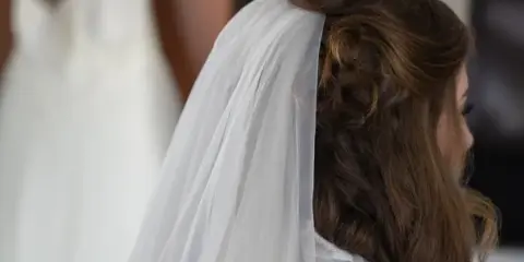 A bride with a styled updo and veil, with a wedding dress hanging in the background.