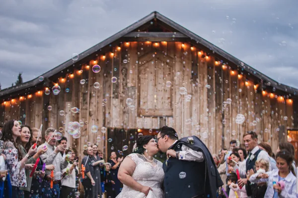 A bride and groom kiss in front of a rustic wooden barn venue while guests blow bubbles.