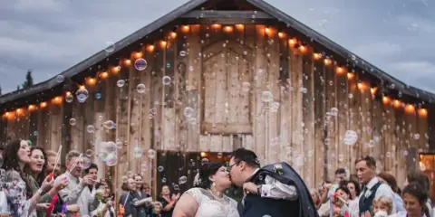 A bride and groom kiss in front of a rustic wooden barn venue while guests blow bubbles.
