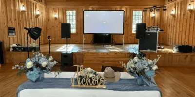 A wedding reception venue with a decorated head table, floral arrangements, and a stage equipped with DJ speakers and a projector screen.