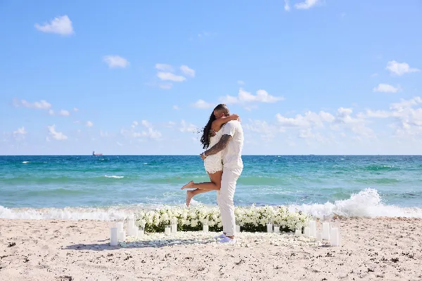 A couple embracing on a beach in front of a floral and candle-staged proposal setup.