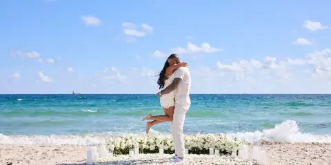 A couple embracing on a beach in front of a floral and candle-staged proposal setup.