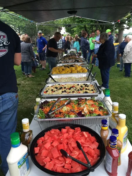 Outdoor catering event with a long table of food including salads and watermelon, surrounded by guests.