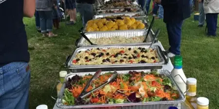 Outdoor catering event with a long table of food including salads and watermelon, surrounded by guests.