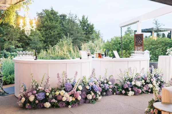 A wedding bar decorated with a lush, sprawling floral arrangement in shades of purple, pink, and white, set against a backdrop of greenery and a chandelier.