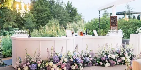 A wedding bar decorated with a lush, sprawling floral arrangement in shades of purple, pink, and white, set against a backdrop of greenery and a chandelier.
