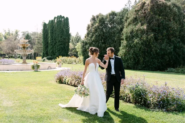 A bride in a white gown and a groom in a black tuxedo walking together in a garden with a fountain in the background.