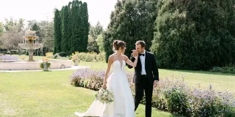 A bride in a white gown and a groom in a black tuxedo walking together in a garden with a fountain in the background.