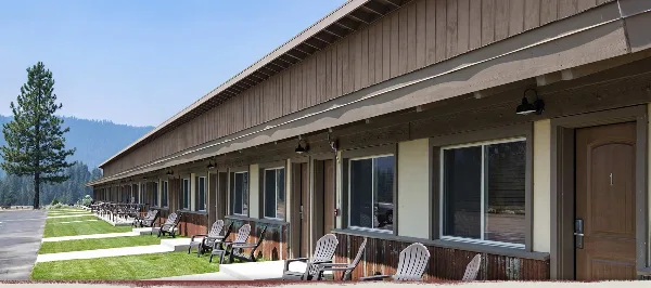 Exterior view of a long, single-story wooden lodge with multiple guest rooms, outdoor Adirondack chairs, and a mountain backdrop.