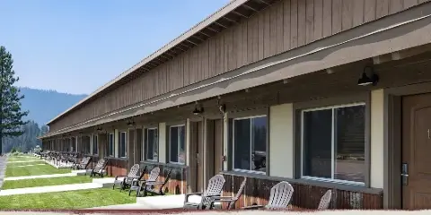 Exterior view of a long, single-story wooden lodge with multiple guest rooms, outdoor Adirondack chairs, and a mountain backdrop.