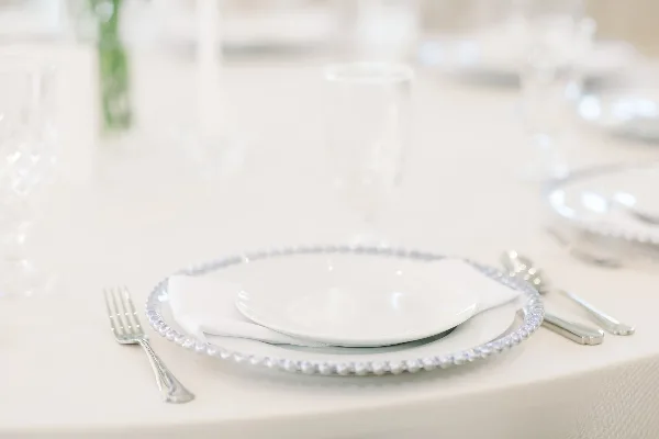 Styled wedding table setting with white plates, beaded charger, silverware, and glassware on a white tablecloth.