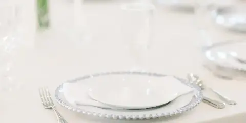 Styled wedding table setting with white plates, beaded charger, silverware, and glassware on a white tablecloth.