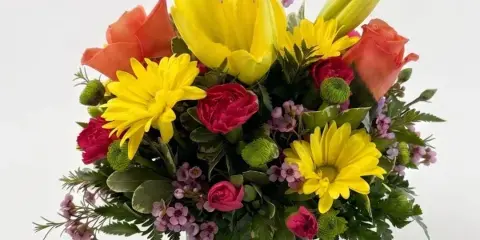 A vibrant floral arrangement featuring yellow lilies, orange roses, and yellow daisies in a clear glass vase.