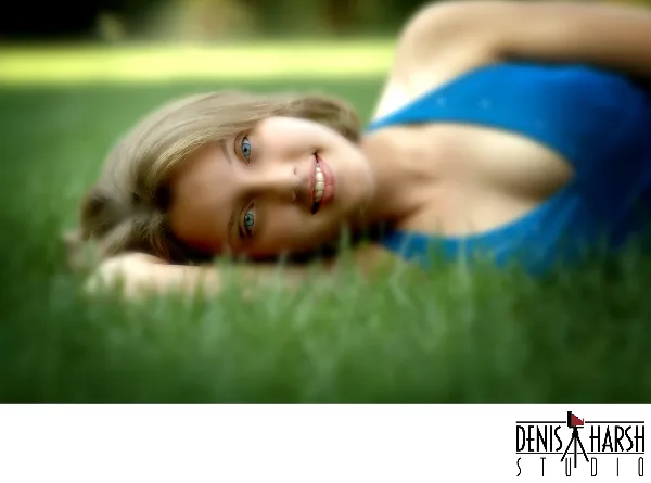 A professional portrait of a smiling young woman with blonde hair lying on green grass, wearing a blue top.