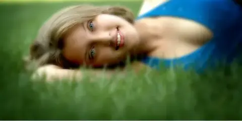 A professional portrait of a smiling young woman with blonde hair lying on green grass, wearing a blue top.