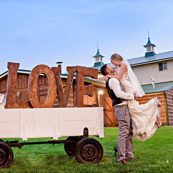 A bride and groom embracing in front of a rustic wooden barn with a metal roof and cupolas, next to a white wagon with large wooden LOVE letters.