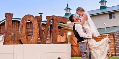 A bride and groom embracing in front of a rustic wooden barn with a metal roof and cupolas, next to a white wagon with large wooden LOVE letters.