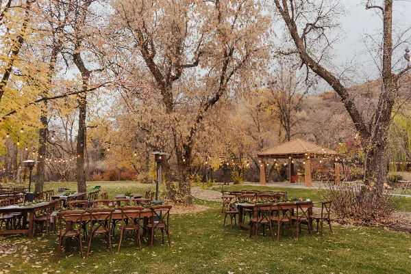 Outdoor event setup with wooden tables, chairs, and string lights in a park setting.