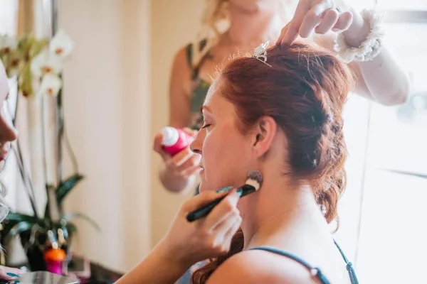 A bride with red hair has her hair styled and makeup applied by artists during wedding preparations.