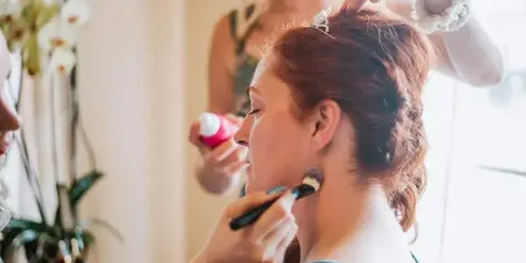A bride with red hair has her hair styled and makeup applied by artists during wedding preparations.
