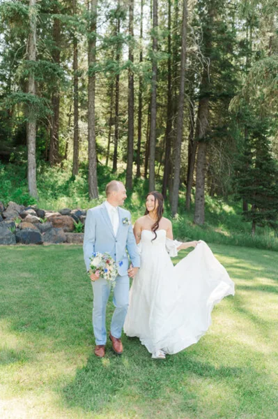 A bride and groom standing in a grassy clearing surrounded by tall pine trees, with the bride holding a bouquet and the groom wearing a light blue suit.