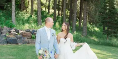 A bride and groom standing in a grassy clearing surrounded by tall pine trees, with the bride holding a bouquet and the groom wearing a light blue suit.