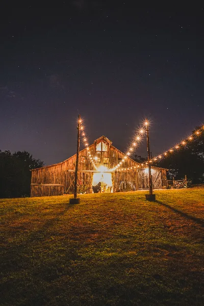 A rustic wooden barn venue illuminated by string lights at night, set on a grassy field.
