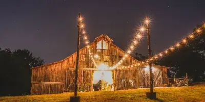 A rustic wooden barn venue illuminated by string lights at night, set on a grassy field.