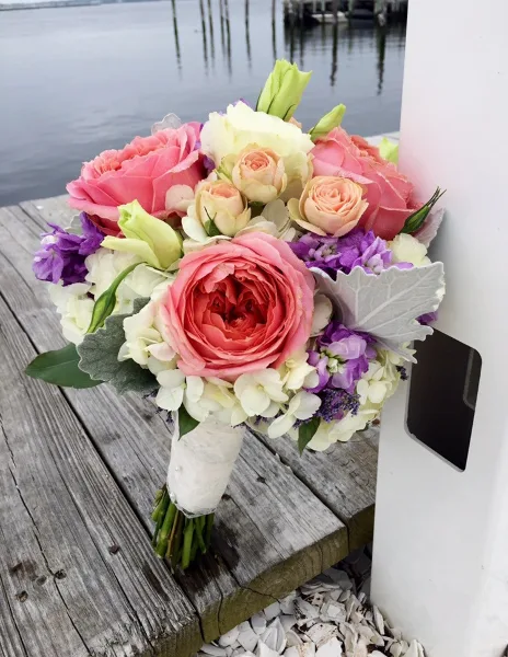 A wedding bouquet with pink and white roses, purple stock, and white hydrangeas resting on a wooden dock.
