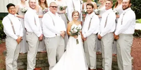 A wedding party including the bride, groom, and wedding party posing outdoors in front of a venue building.