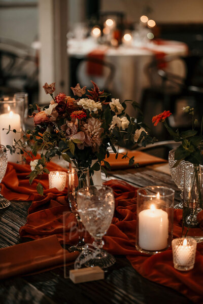 A rustic wedding table setting featuring a floral centerpiece, lit candles, and orange fabric runners.