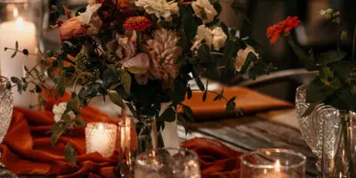 A rustic wedding table setting featuring a floral centerpiece, lit candles, and orange fabric runners.