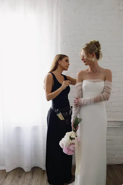 A professional hair and makeup artist adjusting the hair of a bride in a white wedding dress.
