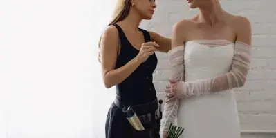 A professional hair and makeup artist adjusting the hair of a bride in a white wedding dress.