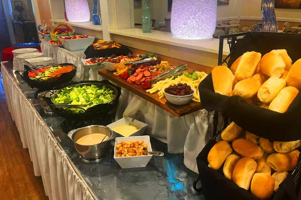 A catering buffet table featuring a salad, charcuterie board, cheese, and baskets of bread rolls.